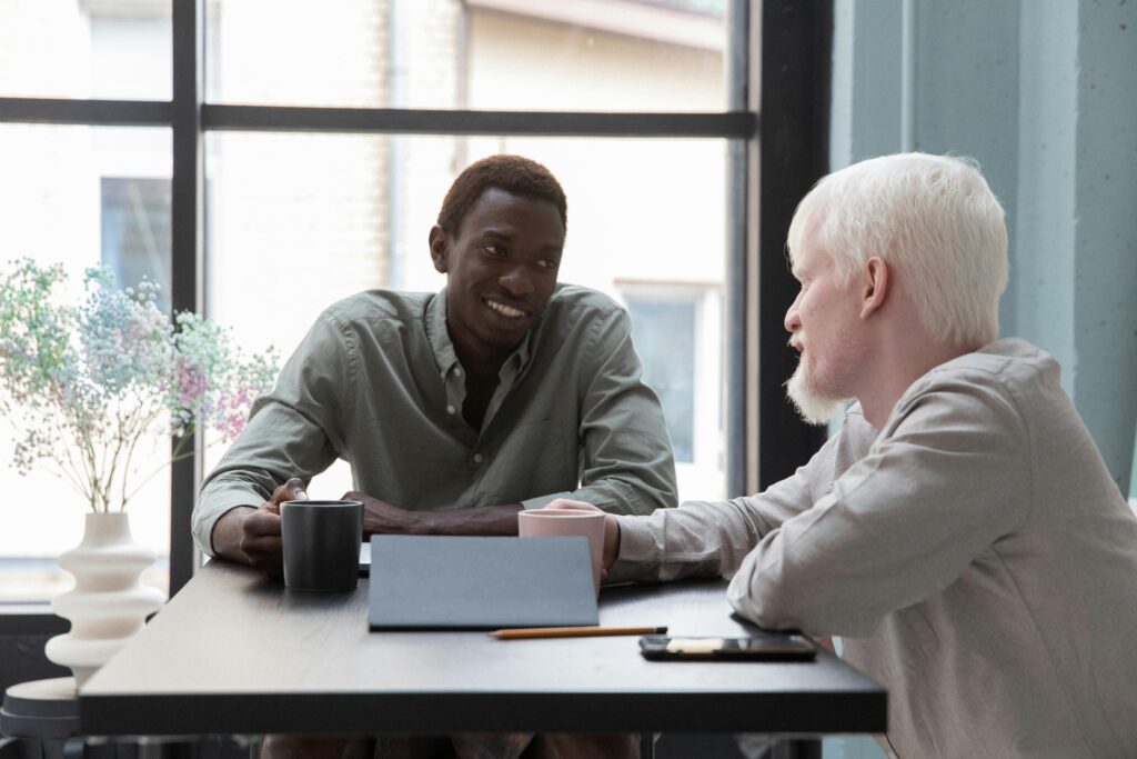 Cheerful African American man sitting at table with albino coworker while drinking coffee and talking about work in modern office