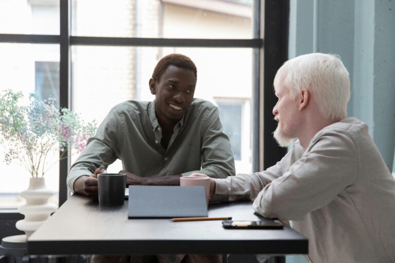 Cheerful African American man sitting at table with albino coworker while drinking coffee and talking about work in modern office